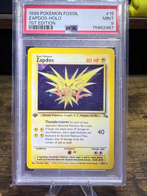 A graded 1999 Pokémon Fossil Zapdos Holo card, PSA 1st Edition Mint 9, sealed in a clear protective plastic slab, set against a dark wooden background.