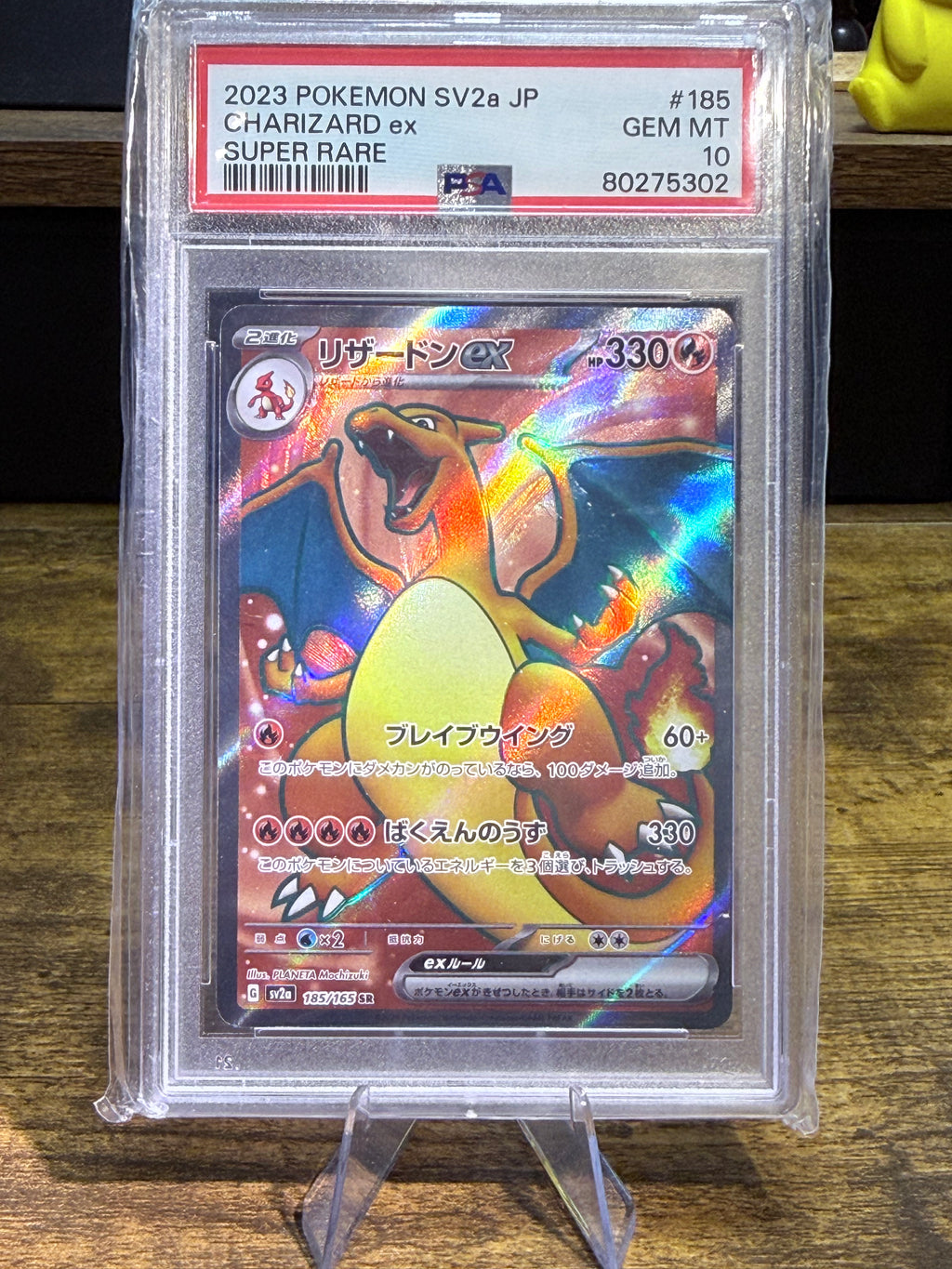 A graded Pokémon Charizard SV2 2a JP trading card inside a clear PSA Gem Mint protective case, displayed on a stand over a wooden surface.