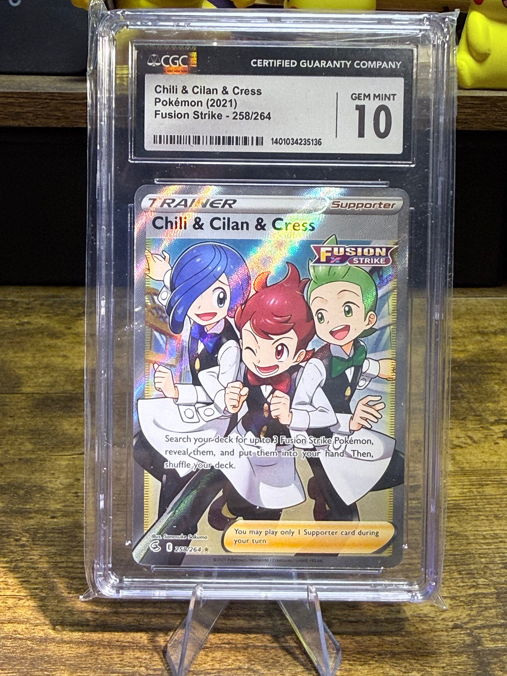 A graded Pokémon trading card in a clear plastic grading case on a small display stand, photographed on a wooden surface. The card is Chili & Cilan & Cress from Fusion Strike, and the top label shows a Gem Mint 10 from CCG.