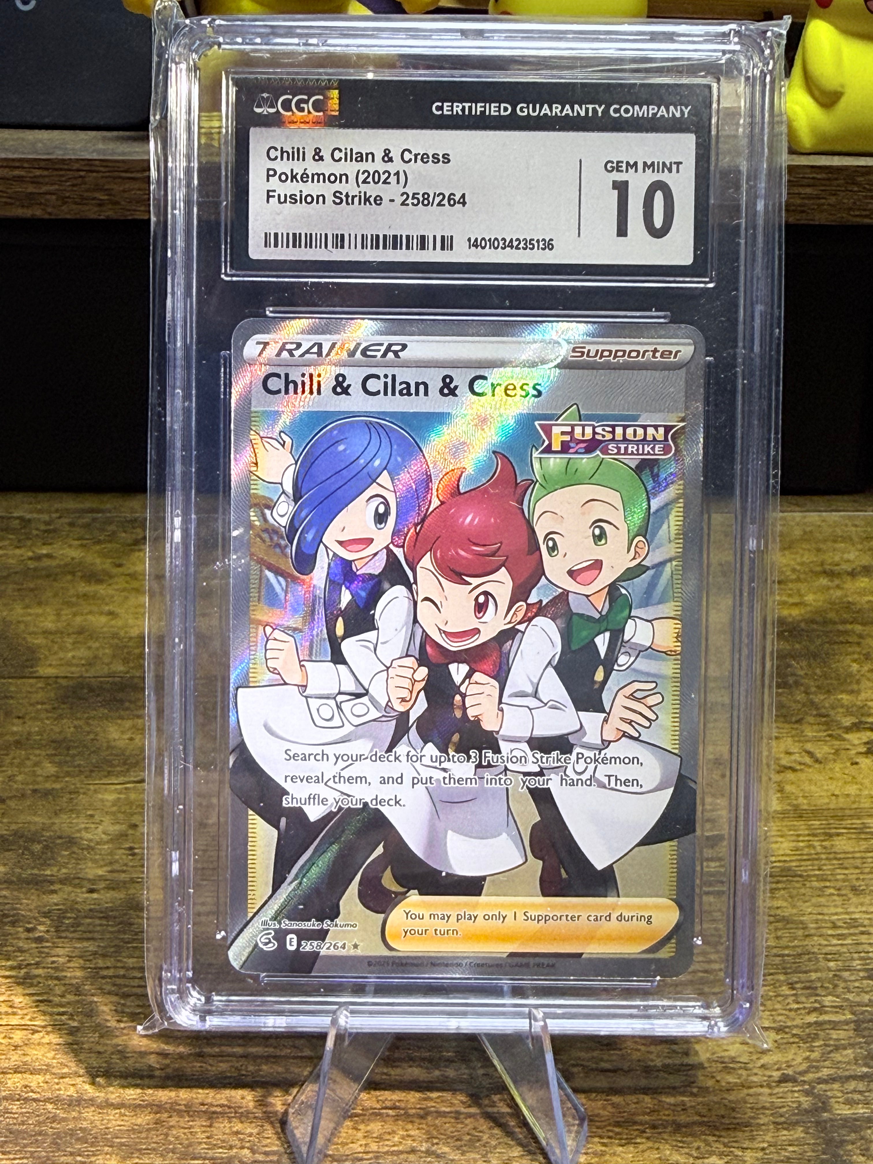 A graded Pokémon trading card in a clear plastic grading case on a small display stand, photographed on a wooden surface. The card is Chili & Cilan & Cress from Fusion Strike, and the top label shows a Gem Mint 10 from CCG.
