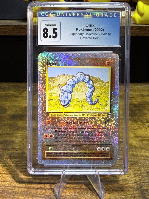 A graded holographic Pokémon card of Onix from the 2002 Legendary Collection, graded 8.5 by CGC, displayed in a clear plastic slab on a stand over a wooden surface.