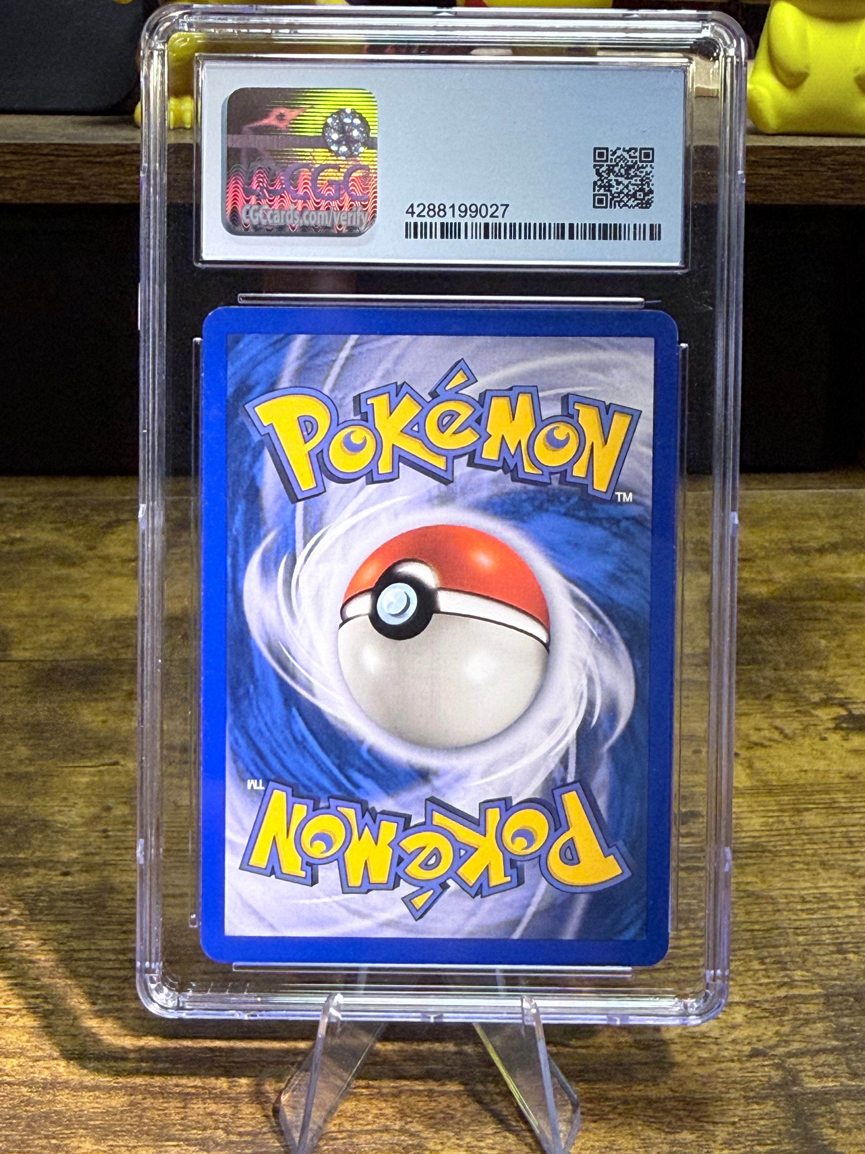 A graded Pokémon card in a clear plastic PSA-style holder, standing on a small clear display stand on a wooden surface. The card has a blue background with a red-and-white Poké Ball illustration and yellow 'Pokémon' lettering, and the grading label is visible at the top.