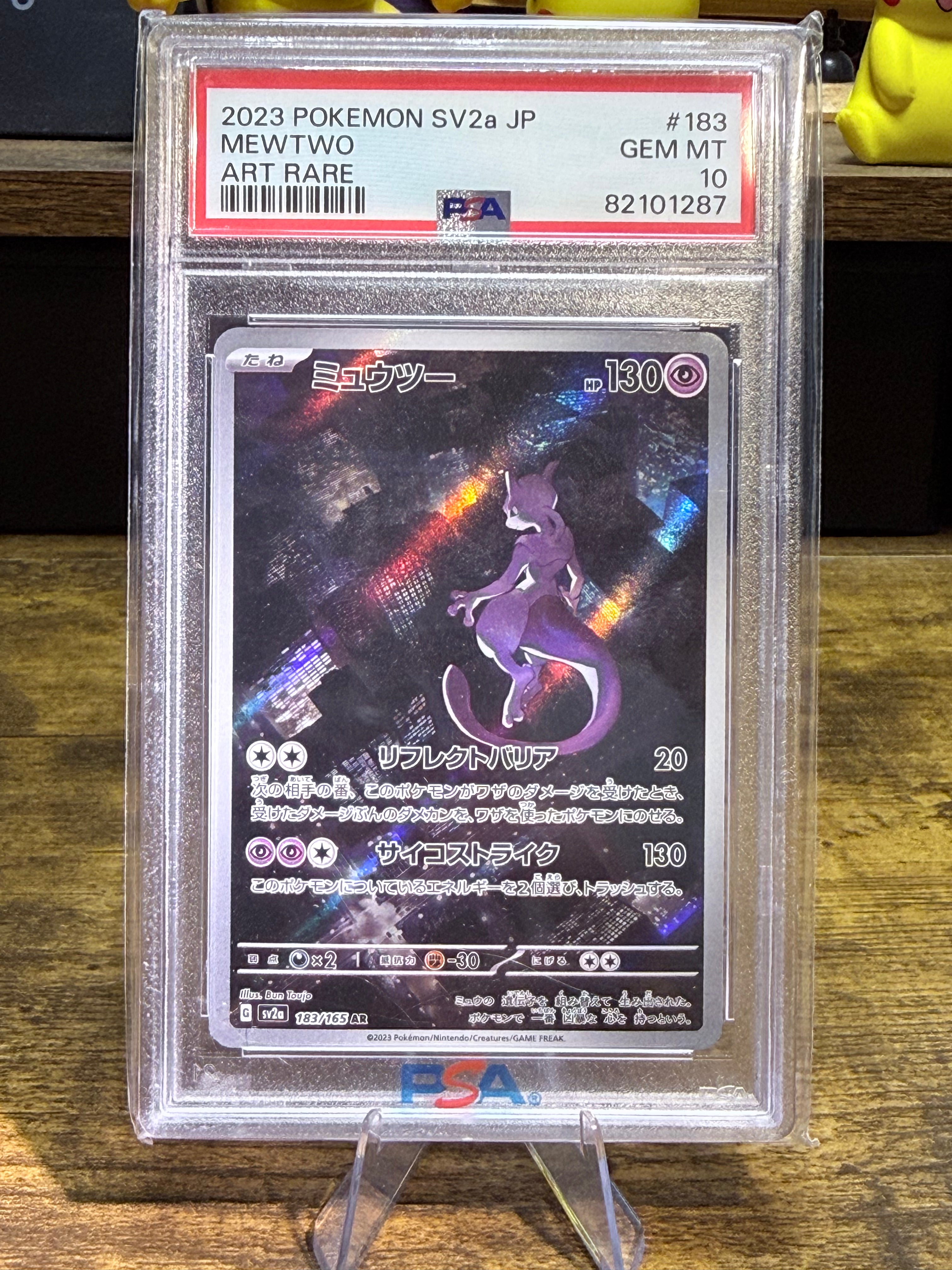 A graded Pokémon card of Mewtwo from 2023, showing the Art Rare version. The card is encapsulated in a PSA grading holder and displayed on a clear stand on a wooden surface.