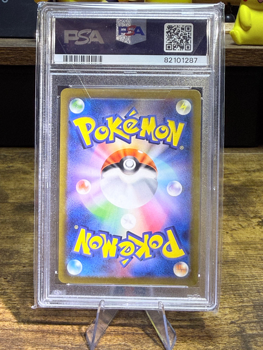 A graded PSA PSA+ Pokémon card in a clear plastic slab, displayed on a clear stand on a wooden surface.